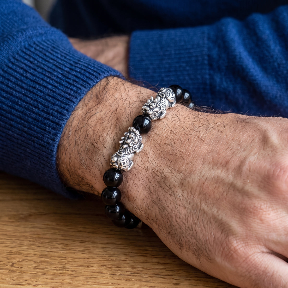 A close-up of a person's wrist wearing a beaded bracelet with black round beads and ornate silver-toned metal links, against a blue sweater and wooden table background.