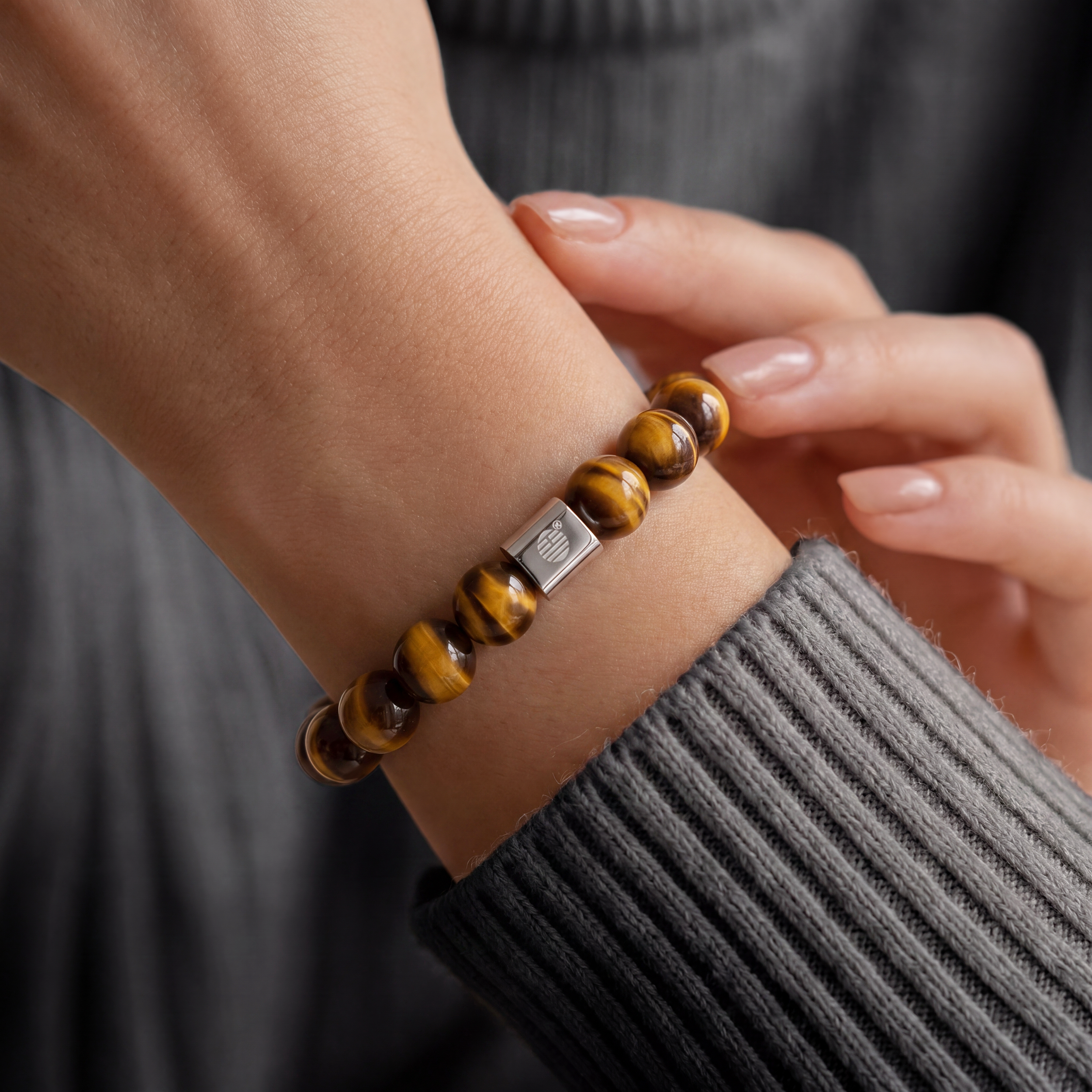 A close-up of a person's wrist wearing a beaded bracelet made of round brown and golden-yellow beads with a silver-toned rectangular accent clasp, paired with a dark gray ribbed sweater.