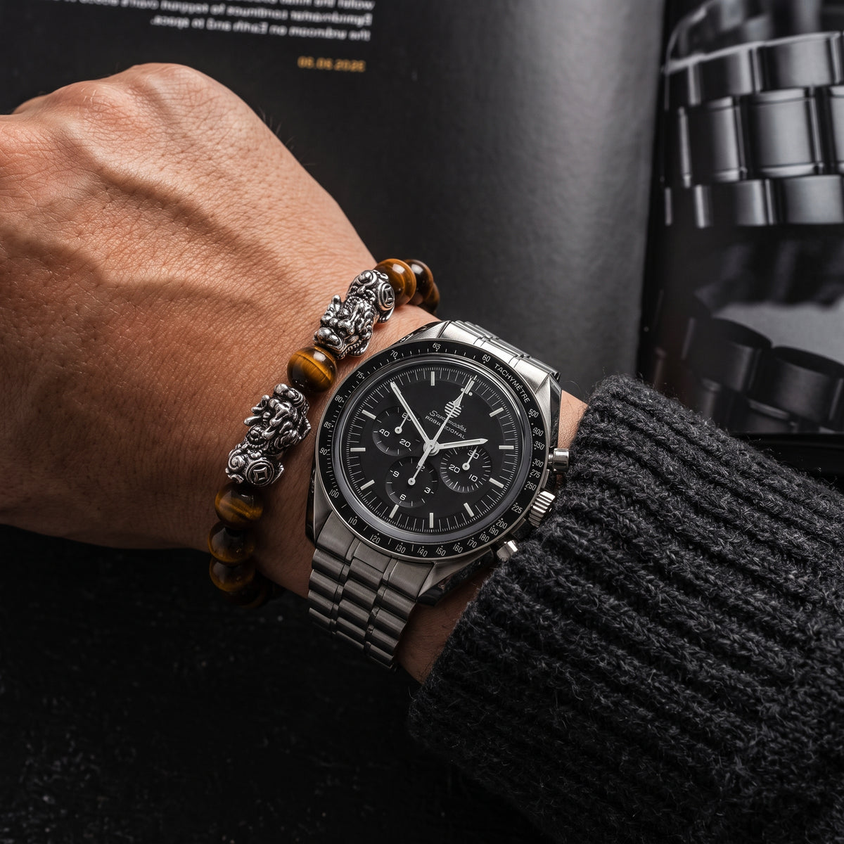 A close-up of a man's wrist wearing a stainless steel chronograph watch with a black dial and bracelet, along with a silver beaded bracelet and a dark knit sweater sleeve.