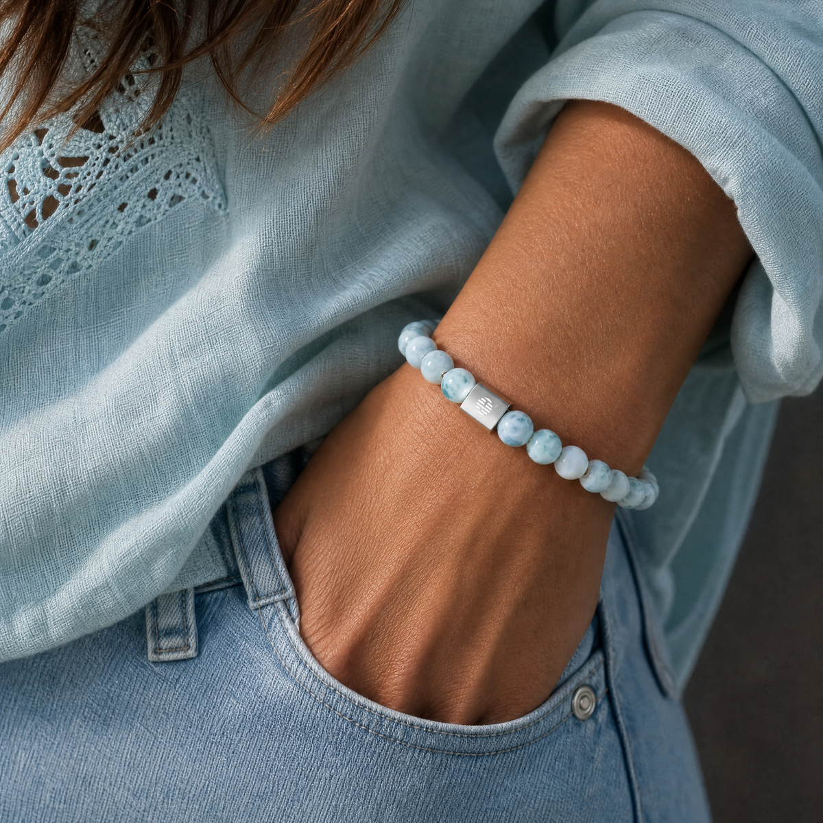 A close-up of a person's wrist wearing a light blue beaded bracelet with a small silver charm, paired with a light blue blouse and jeans.