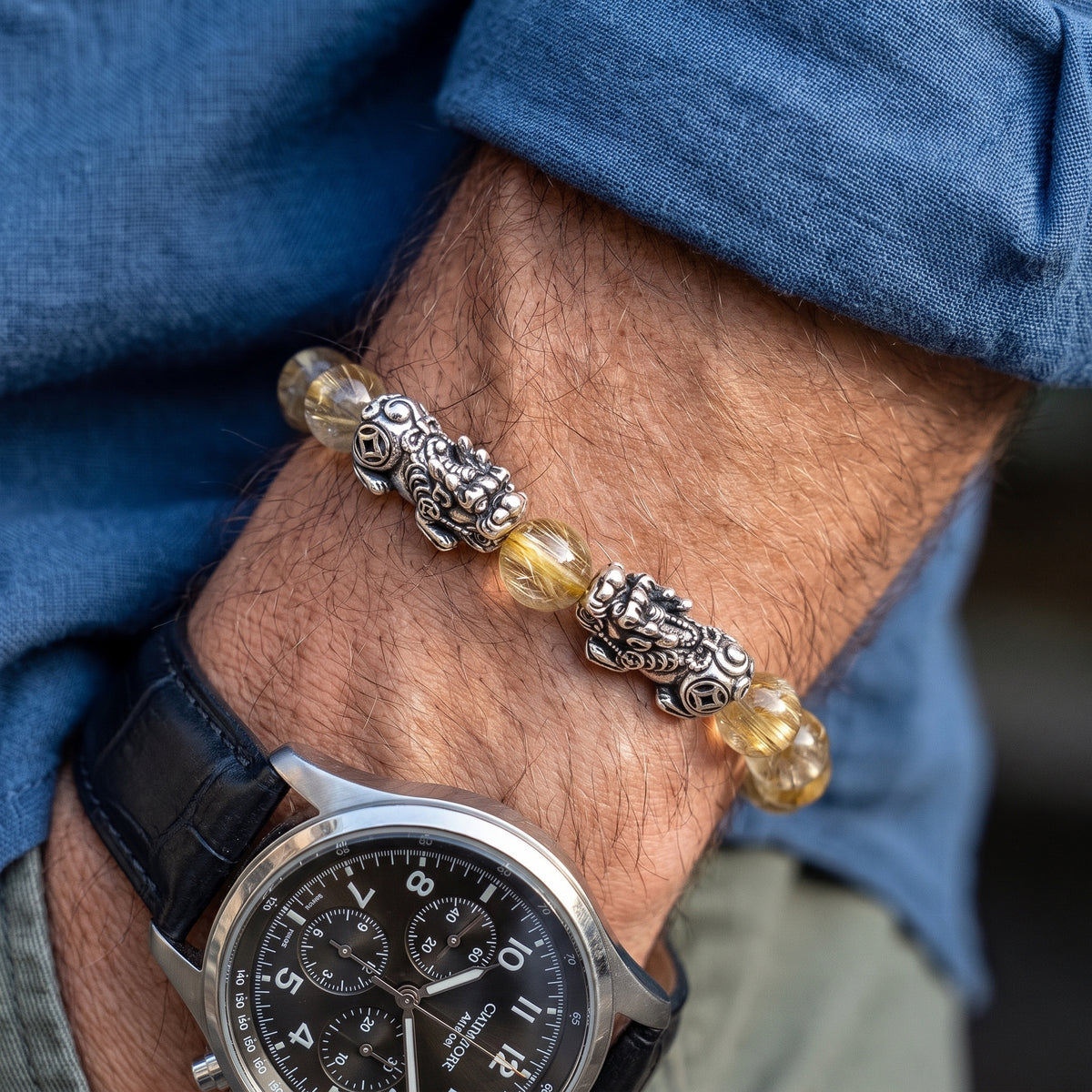 A close-up of a man's wrist wearing a silver-toned beaded bracelet with lion head charms and translucent yellow beads, layered over a black leather watch strap and blue denim shirt.