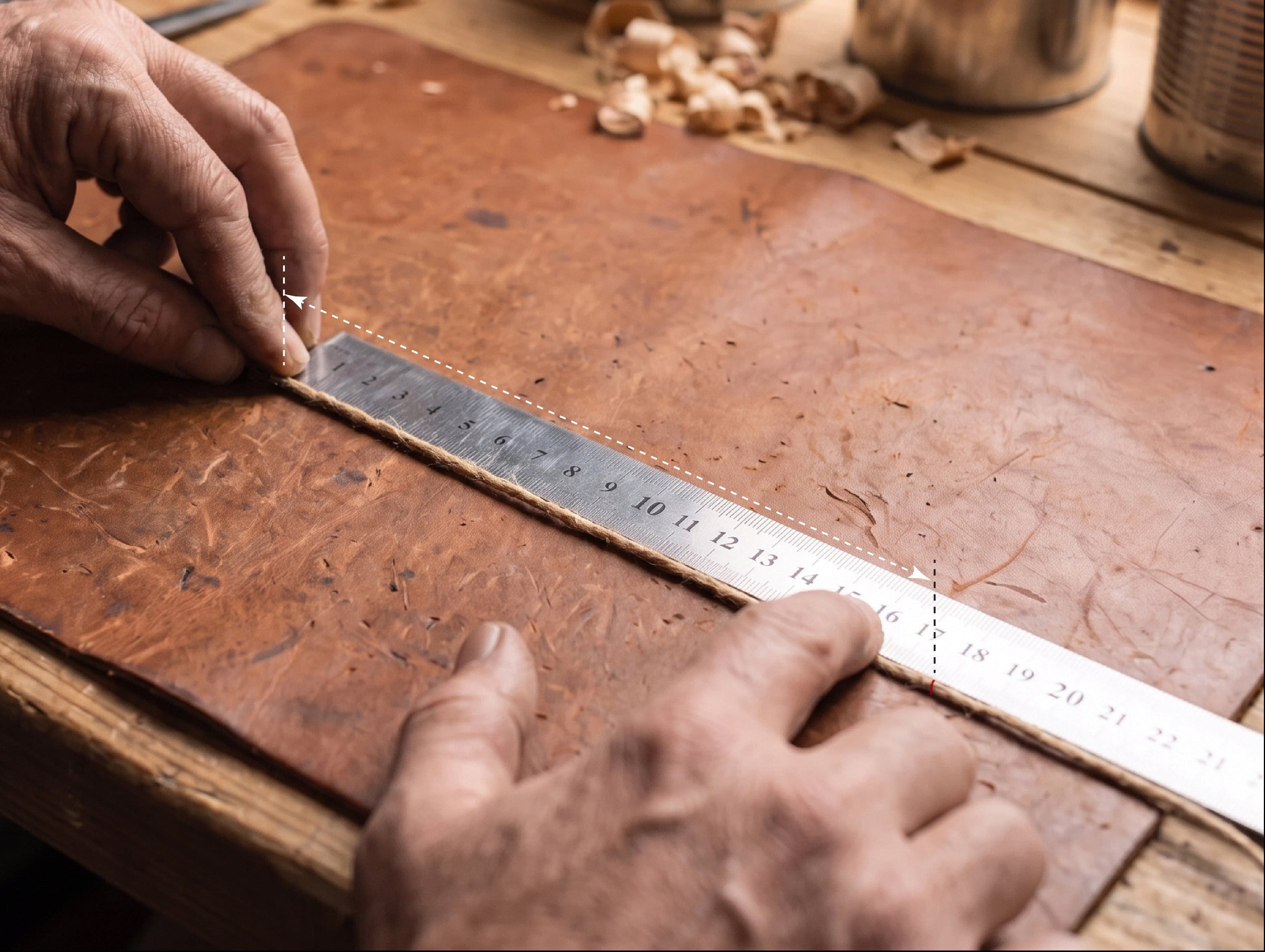 A close-up of a craftsman's hands using a metal ruler to measure a brown leather board on a wooden workbench, with wood shavings and metal cans in the background.