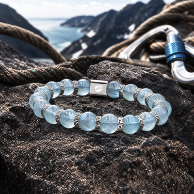 A beaded stretch bracelet made of light blue translucent gemstone beads with silver Greek key spacer rings and a central silver square charm, displayed on a dark rock with a mountain and ocean background.