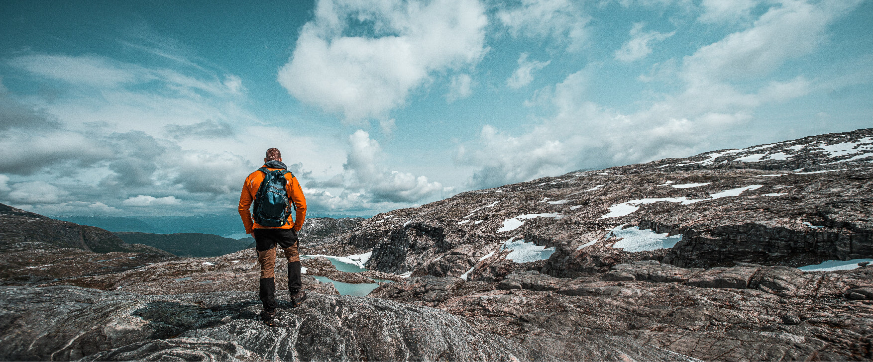 A hiker in an orange jacket and black backpack stands on rocky mountain ground, looking out over a vast landscape with patches of snow, distant hills, and a blue sky filled with white clouds.