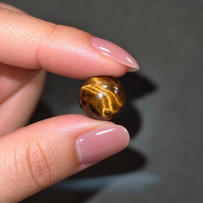 A close-up of a hand with pink manicured nails holding a small, polished golden-brown tiger's eye bead against a dark gray background.