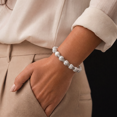 A close-up of a person's hand and wrist wearing a beaded bracelet made of white beads with gray marbled patterns, styled with a cream blouse and beige trousers.