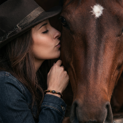 A close-up lifestyle shot of a woman in a dark brown cowboy hat and denim jacket kissing a brown horse on the nose. She is wearing a beaded bracelet on her wrist.